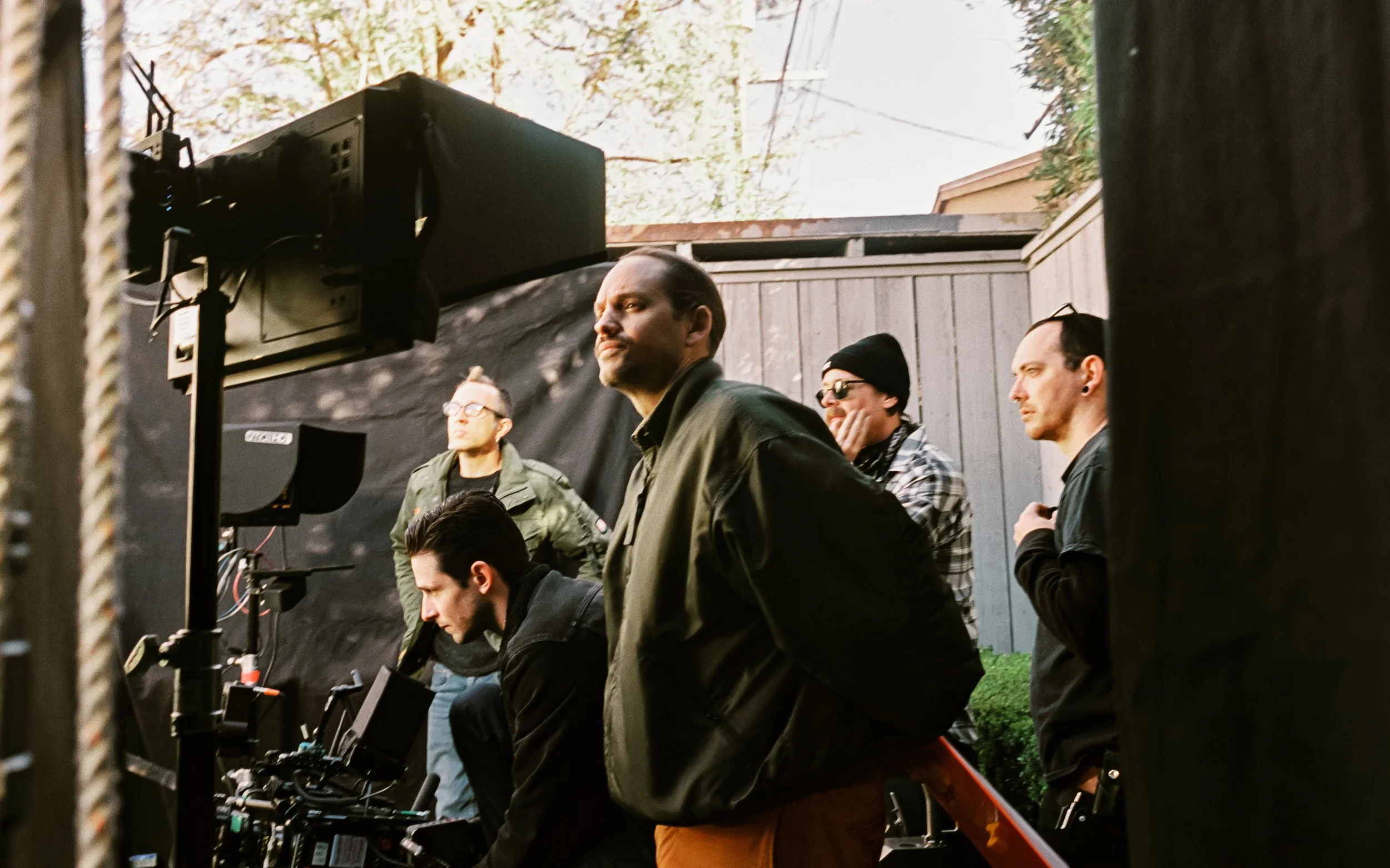 Film crew working on a shoot outdoors with camera equipment and crew members near a wooden fence and trees.