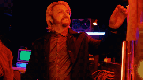 A man with blonde hair and a beard, wearing a dark jacket and shirt, drawing or writing on a vertical surface in a dimly lit room with colorful lights and electronic equipment.