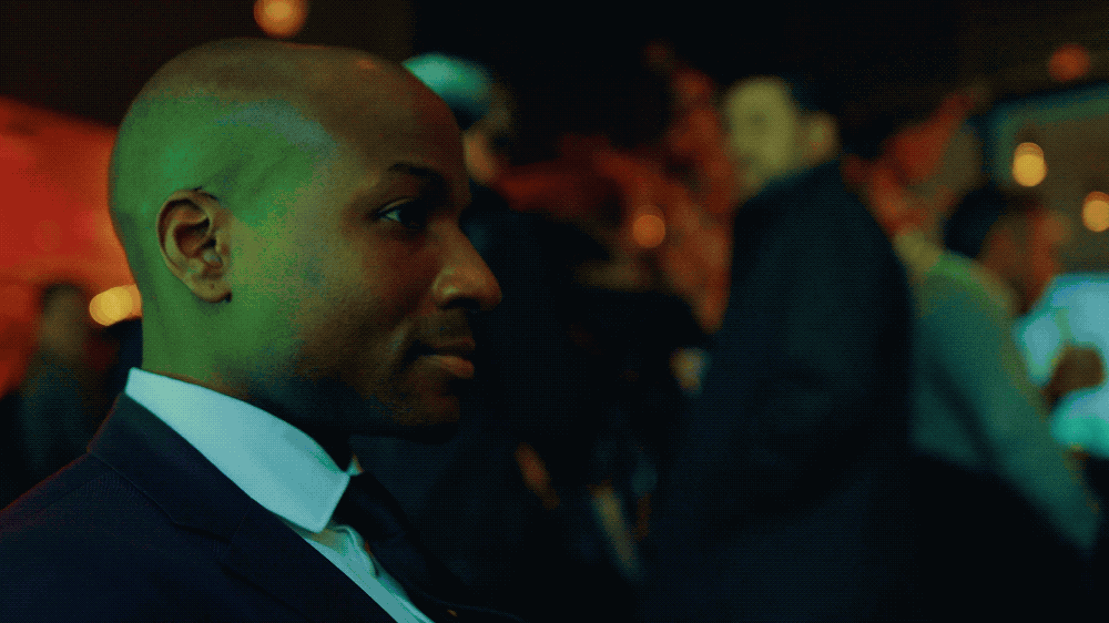 Side profile of a man in a suit attending an event, seated among other people in a dimly lit environment with colorful lights.