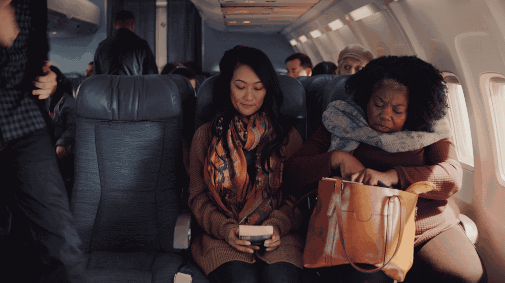 Two women sitting on an airplane, one looking at her phone and the other resting her head on her arms, with other passengers seated and walking in the background.