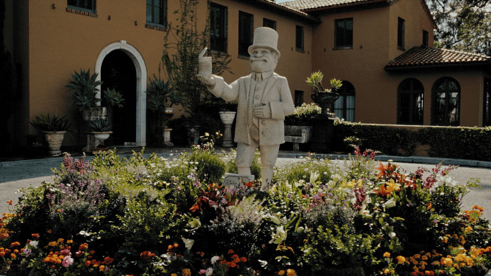 A white statue of a man in a suit and top hat holding a cup, surrounded by colorful flowers in a garden courtyard, with a yellow building in the background.