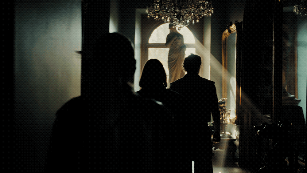 A darkly lit interior with four people walking toward a window, where sunlight streams in and a white statue is visible, with ornate decor and a chandelier overhead.