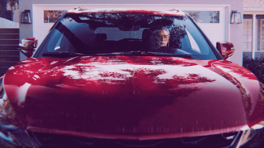 A person with glasses sitting inside a shiny red car, viewed from the front through the windshield, parked in front of a house with a white garage door.