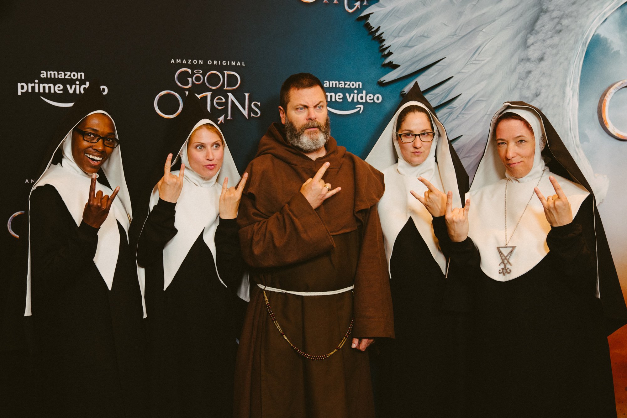 Group of five people dressed as nuns and monks, posing with rock hand gestures in front of a promotional backdrop for Amazon original movie 'Good Omens'.