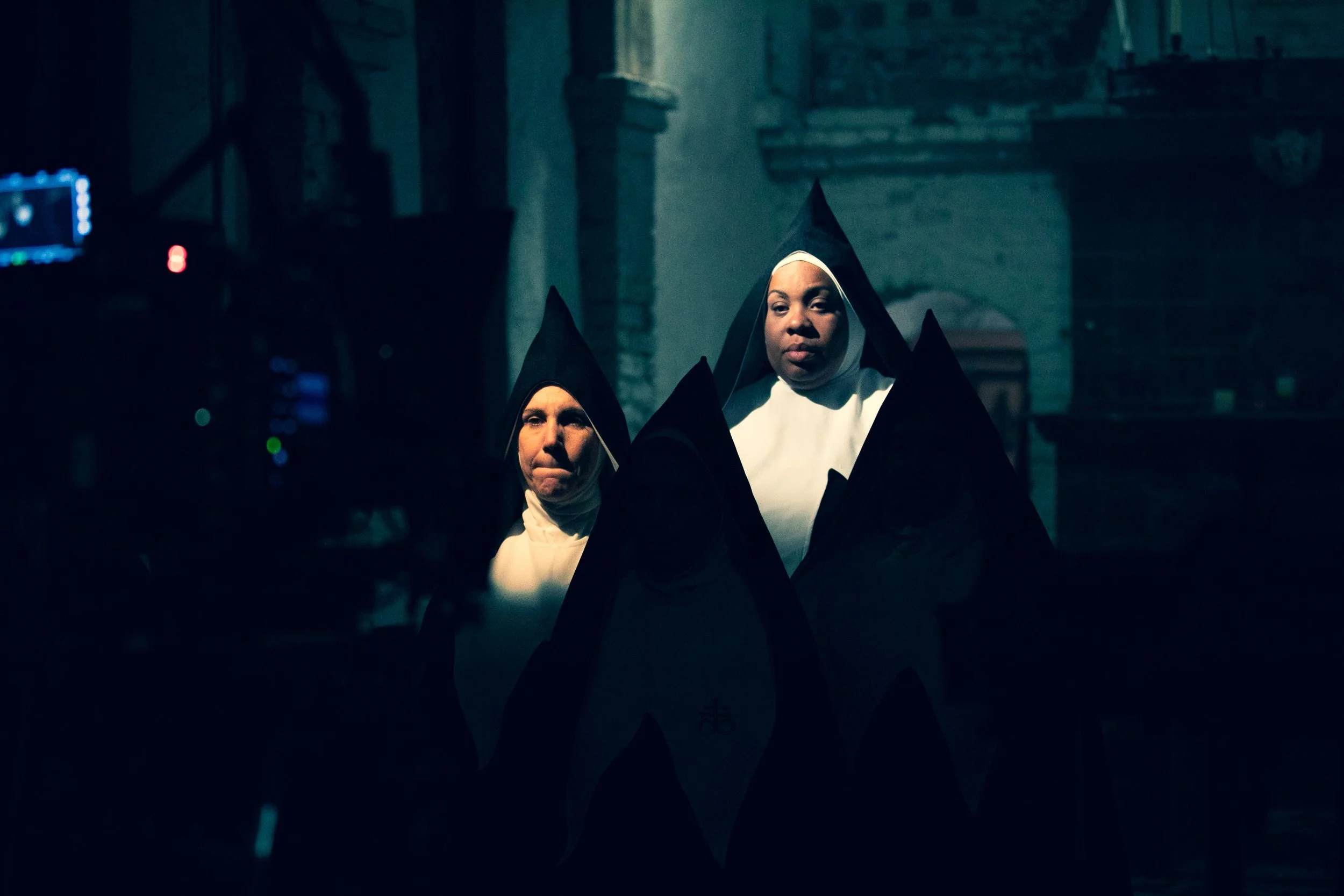 Two women dressed as nuns, wearing black and white habits, standing in a dark, indoor setting with a dimly lit background.Good Omens Amazon Hal Kirkland Director Chattering Order of St Beryl. 
