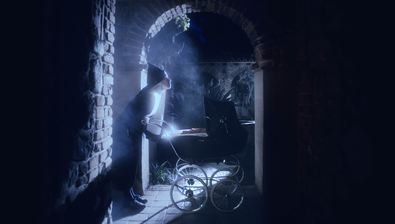 A woman dressed as a nun is standing next to a vintage baby carriage in a dark, arched brick tunnel, with light streaming in from an opening behind her. Good Omens Amazon Hal Kirkland