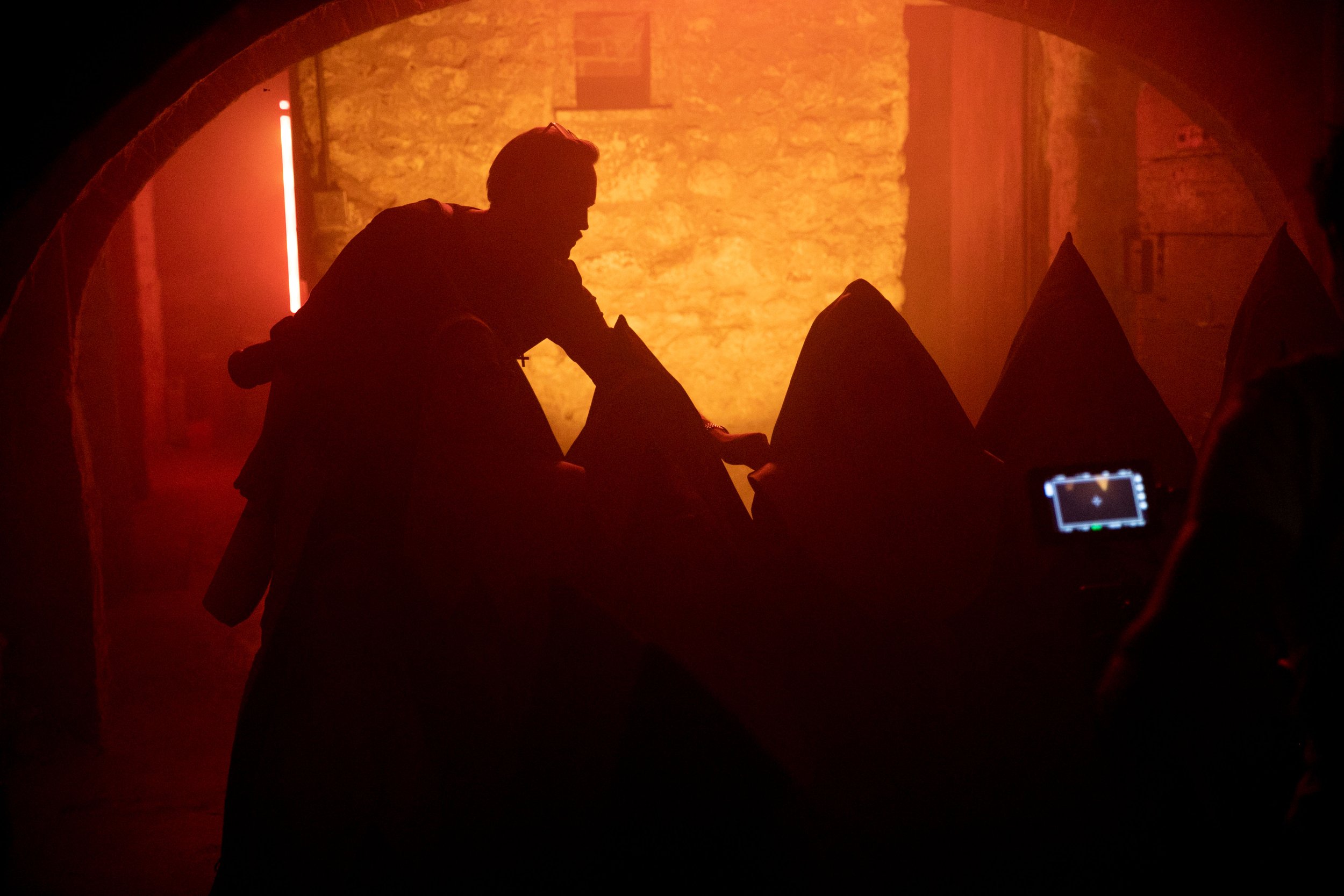 Silhouette of a person in a dimly lit environment with a red hue, appearing to interact with a series of cone-shaped objects, while another person takes a photo or video with a device, in front of a brick wall. Good Omens Amazon Hal Kirkland