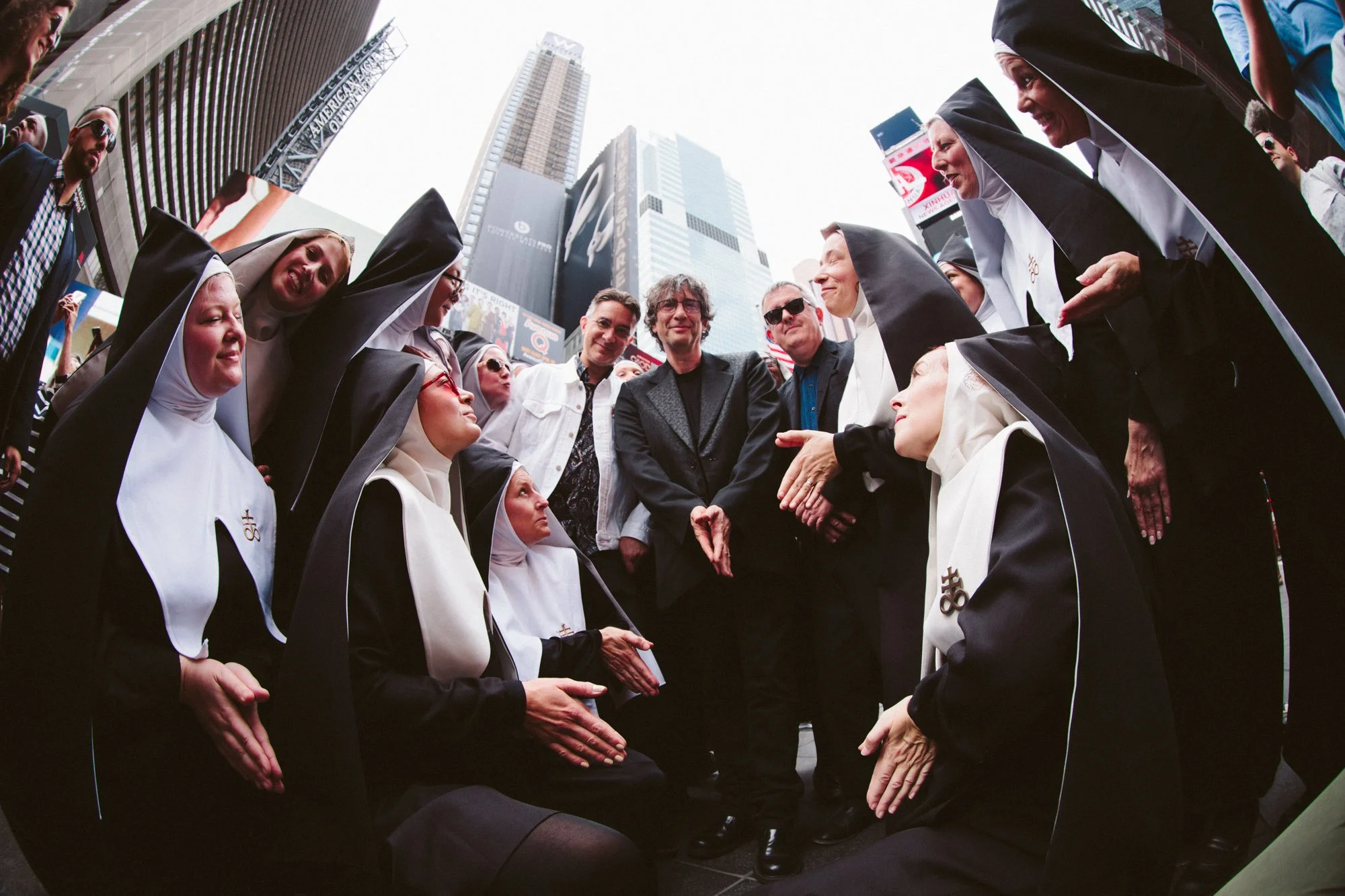 Group of nuns and three men, two of whom are seated and one standing, in Times Square, New York City, with tall buildings and electronic billboards in the background.