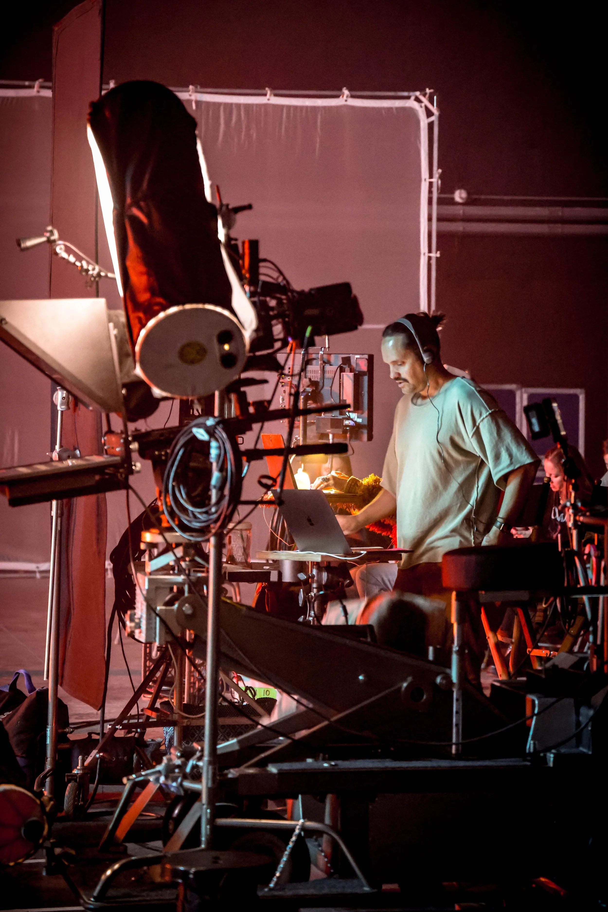 A man wearing a headset working on a laptop at a cluttered table with cameras and equipment, possibly in a studio or behind the scenes of a film or music video production.