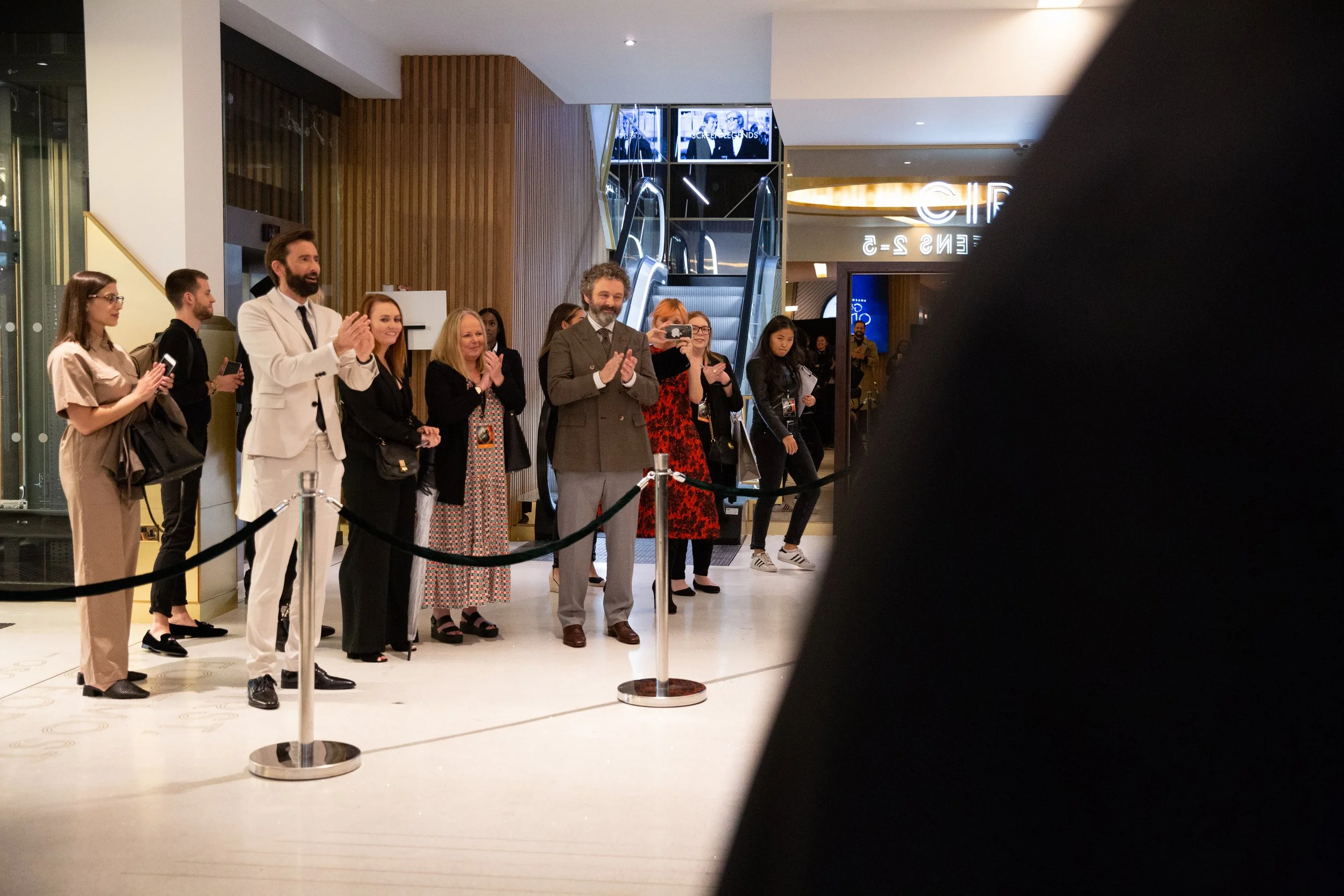 People standing and clapping in a line inside a shopping mall or a lobby, with some taking photos or videos, in front of escalators and a bright sign.