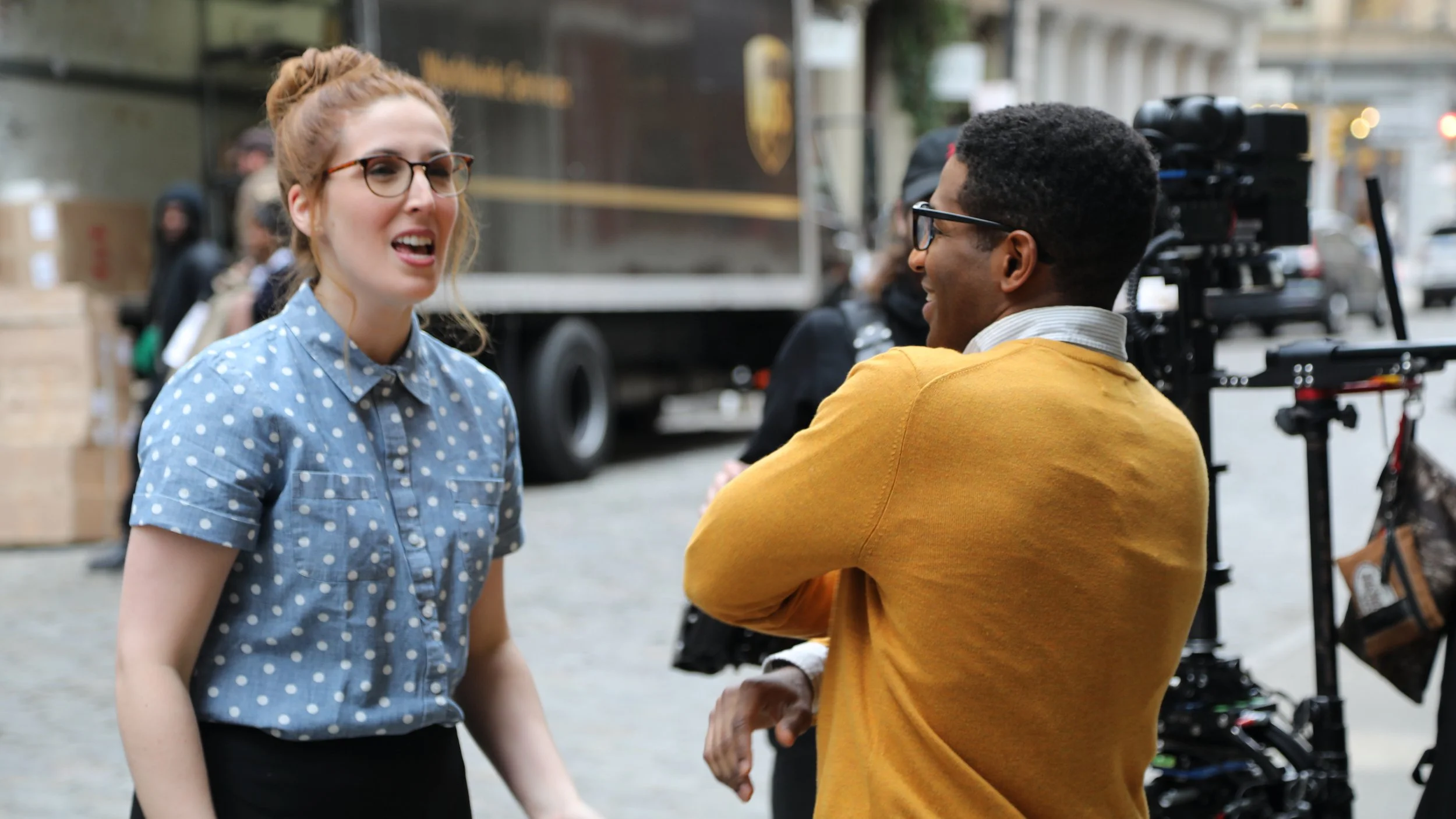 A woman in glasses and a blue polka-dot shirt explains something to a man in glasses and a yellow sweater, who is standing near a camera on a tripod outdoors in an urban setting.