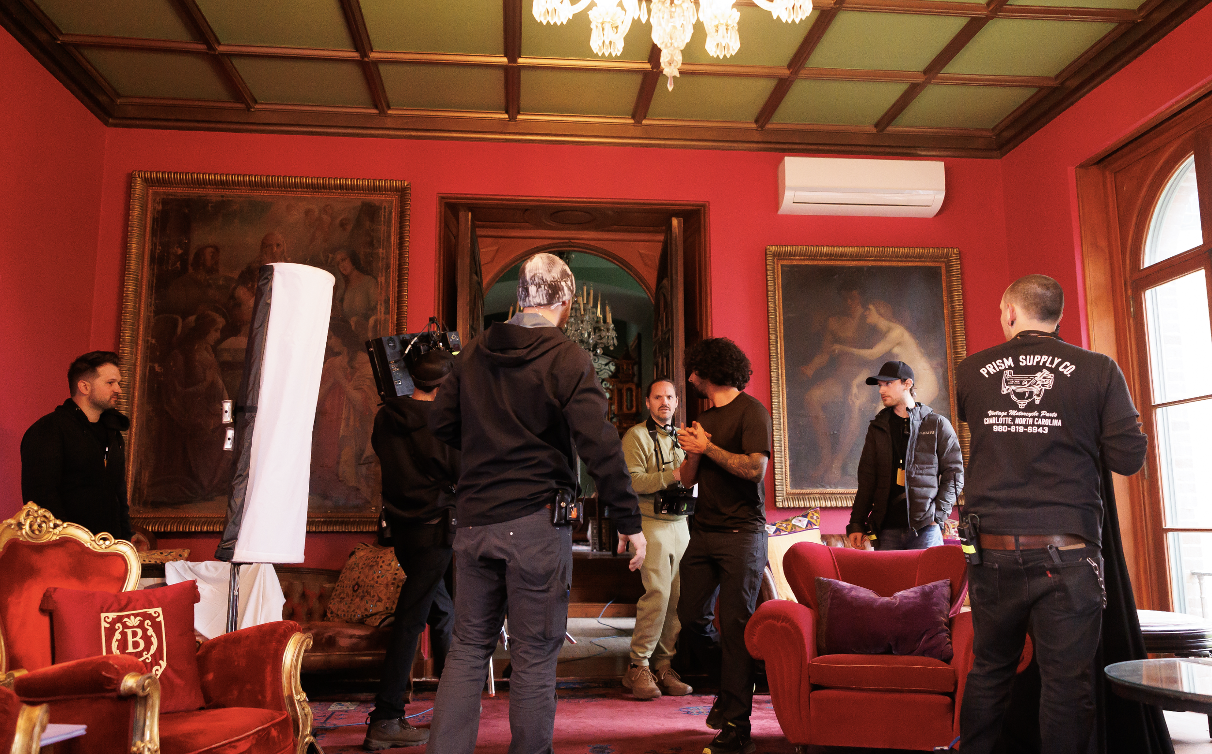 A film crew setting up equipment inside a richly decorated room with red walls, antique furniture, oil paintings, and large windows, possibly filming a scene.