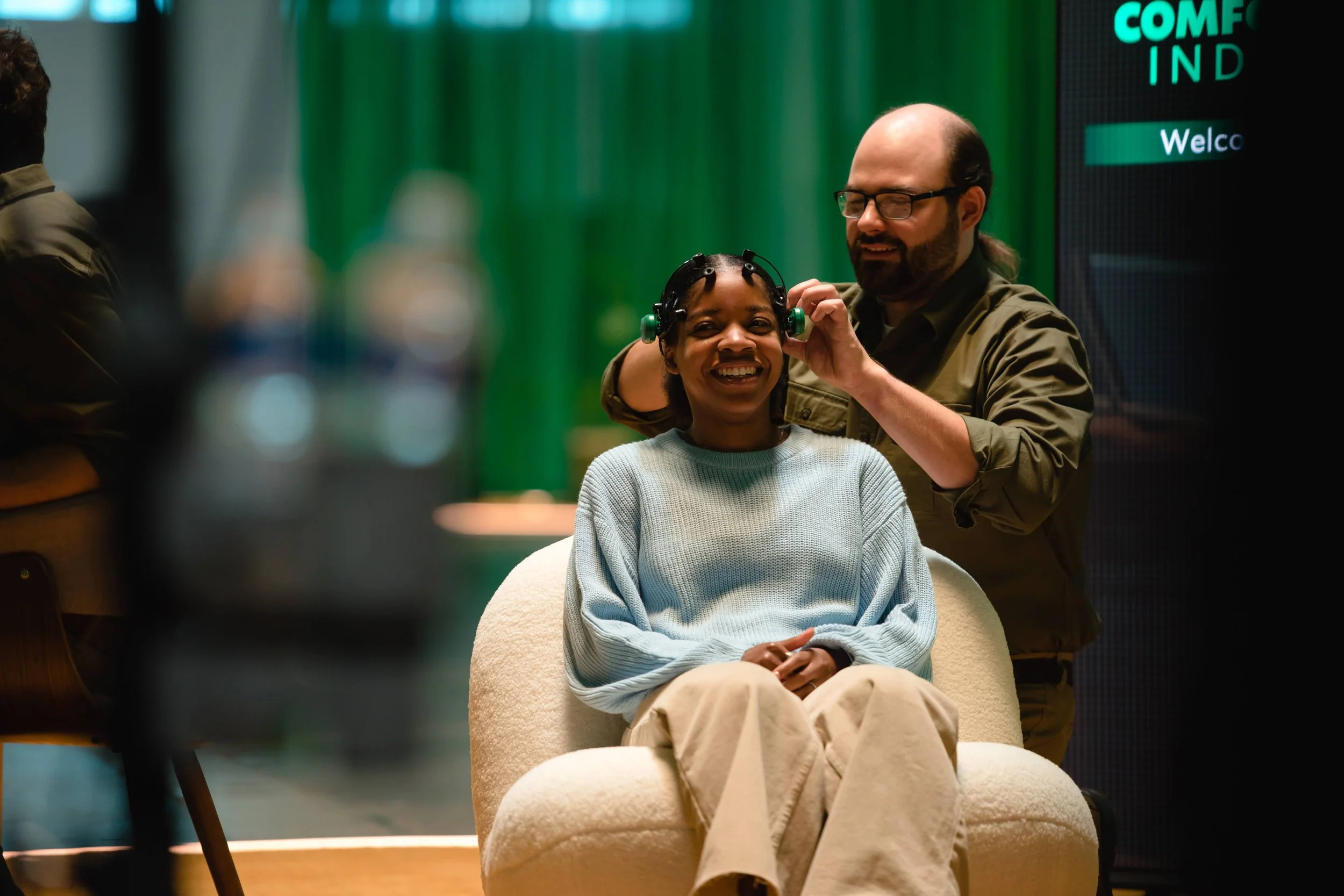 A woman is sitting in a beige chair, smiling, while a man with glasses and a beard assists her with hearing aids in an indoor setting with green background decor.