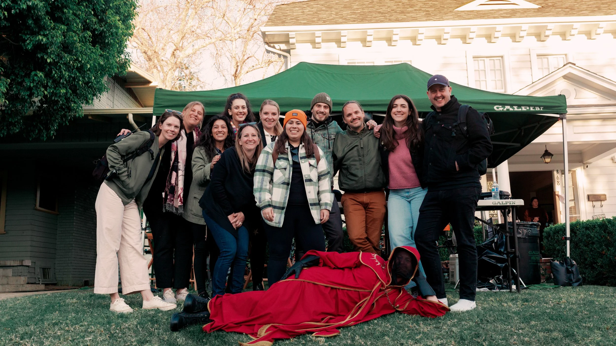 Group of people posing for a photo outdoors, with a person dressed as Santa lying on the grass in front, under a green canopy, in front of a house.