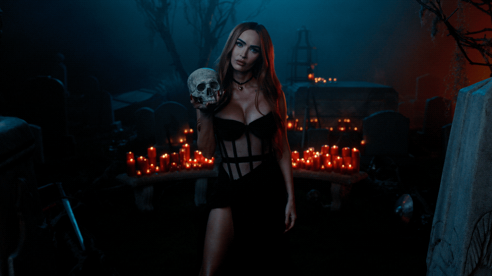A woman with long red hair wearing black lingerie holding a skull in a dark, foggy graveyard decorated with red candles.