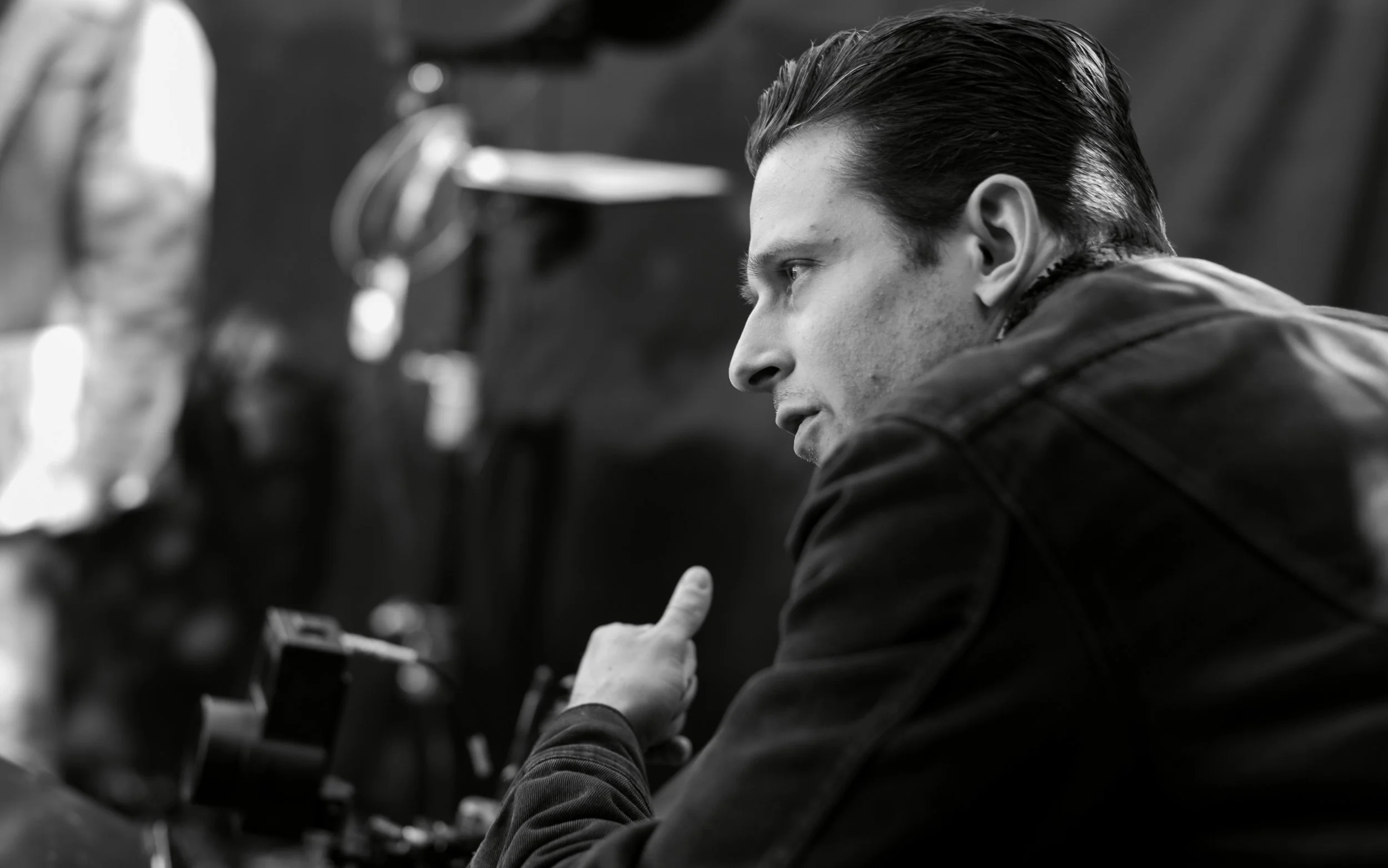 Black and white photo of a young man with slicked-back hair looking intently at something off-camera, with workshop tools blurred in the background.