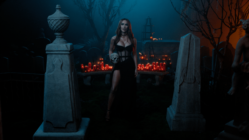 A woman in a black dress holding a skull stands in a dark, eerie cemetery decorated with numerous red candles, stone tombstones, and leafless trees.