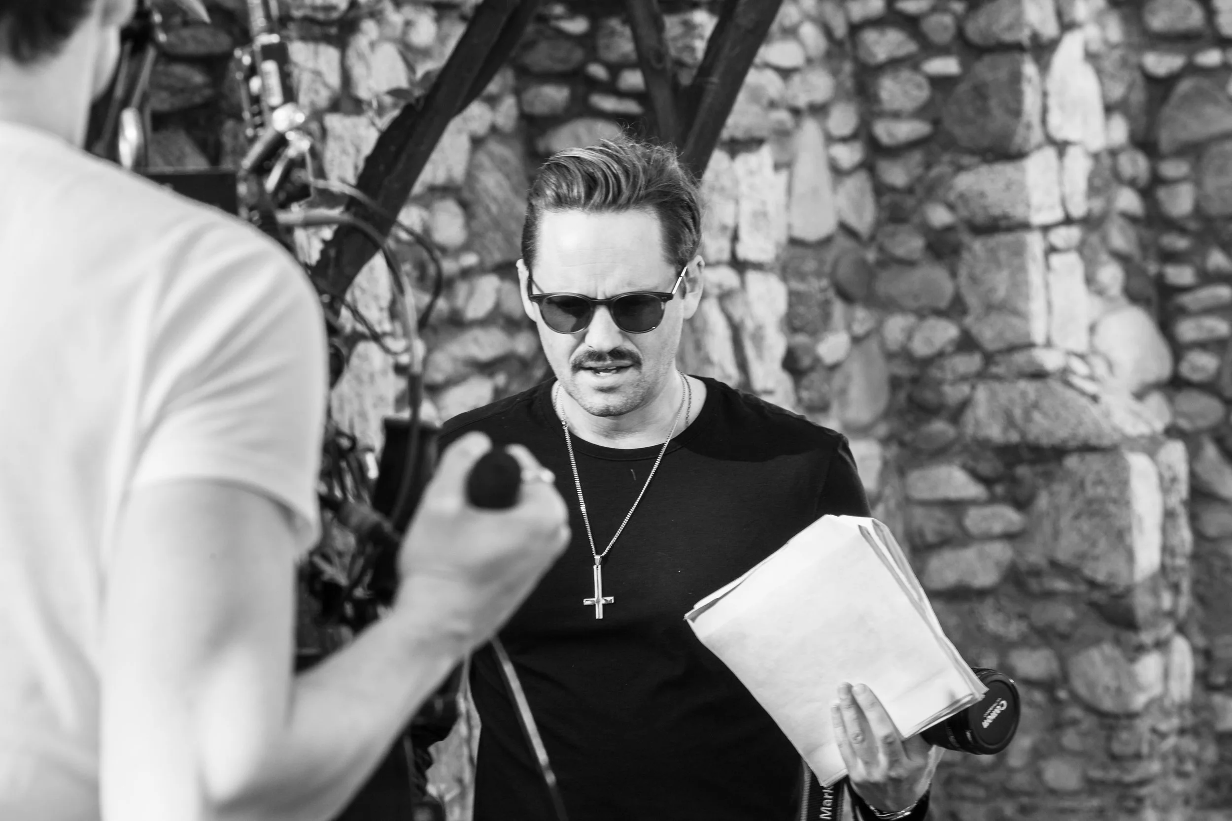 A man wearing sunglasses, a black shirt, and a cross necklace holding a script, is being filmed outdoors with a stone wall in the background. Good Omens Amazon Hal Kirkland Director Chattering Order of St Beryl. 