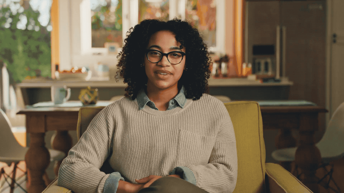 A woman with curly hair and glasses sitting in a comfortable chair in a cozy, well-lit kitchen or living space.