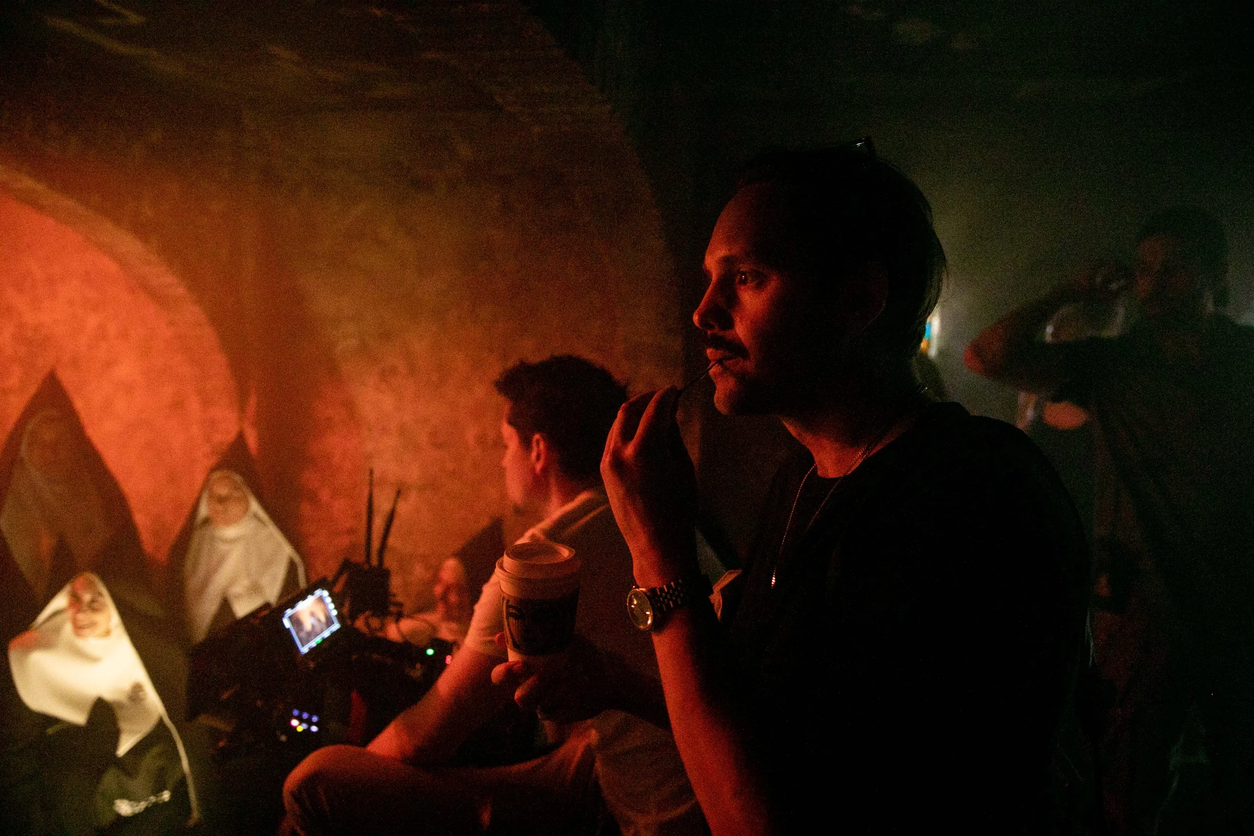 A woman in a dark room holding a coffee cup, with a group of other people around what appears to be a DJ or production setup. Good Omens Amazon Hal Kirkland