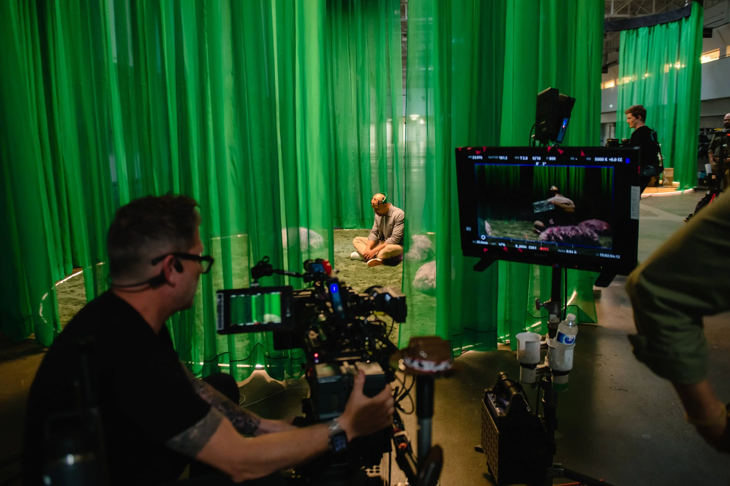 Filming scene with a person sitting in front of a green curtain while a camera crew records with professional equipment.