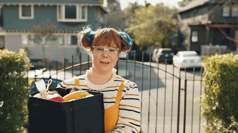 A woman with glasses and colorful hair in pigtails stands outdoors near a black fence, holding a reusable shopping bag filled with groceries, in a residential neighborhood on a sunny day.