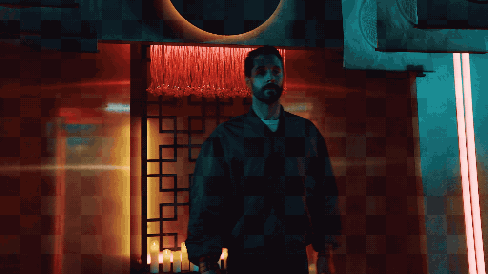 A man with dark hair and facial hair wearing a dark jacket standing in a dimly lit room with colorful neon lights and candles.