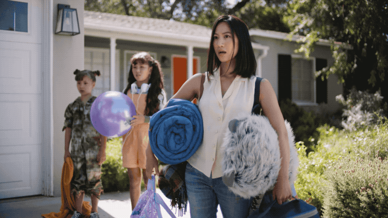 Three children and a woman standing outside a house with a garage. The woman is holding a rolled-up blanket and a furry pillow or stuffed animal, while the children hold balloons and bags. The scene appears to be during daytime.