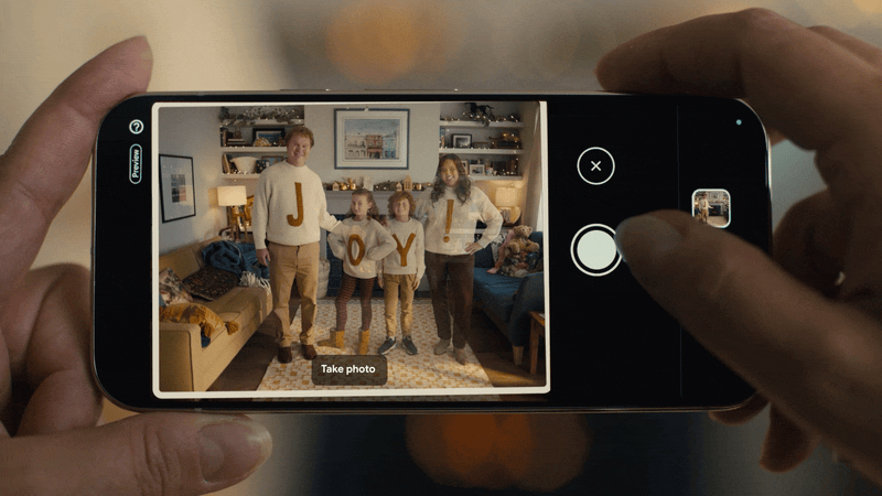A person is taking a photo of a family in a living room, with the family holding up letters spelling 'JOY' on their sweaters.