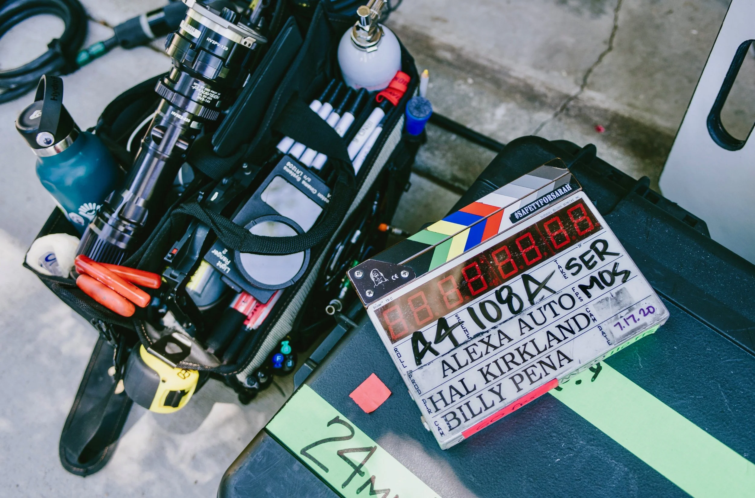 Clapperboard with event details placed on a black case, and a tool bag containing bicycle components, tools, and accessories on a concrete surface.