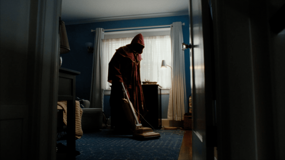 Person in a hooded jacket vacuuming a blue-carpeted room with natural light coming through a window with white curtains.