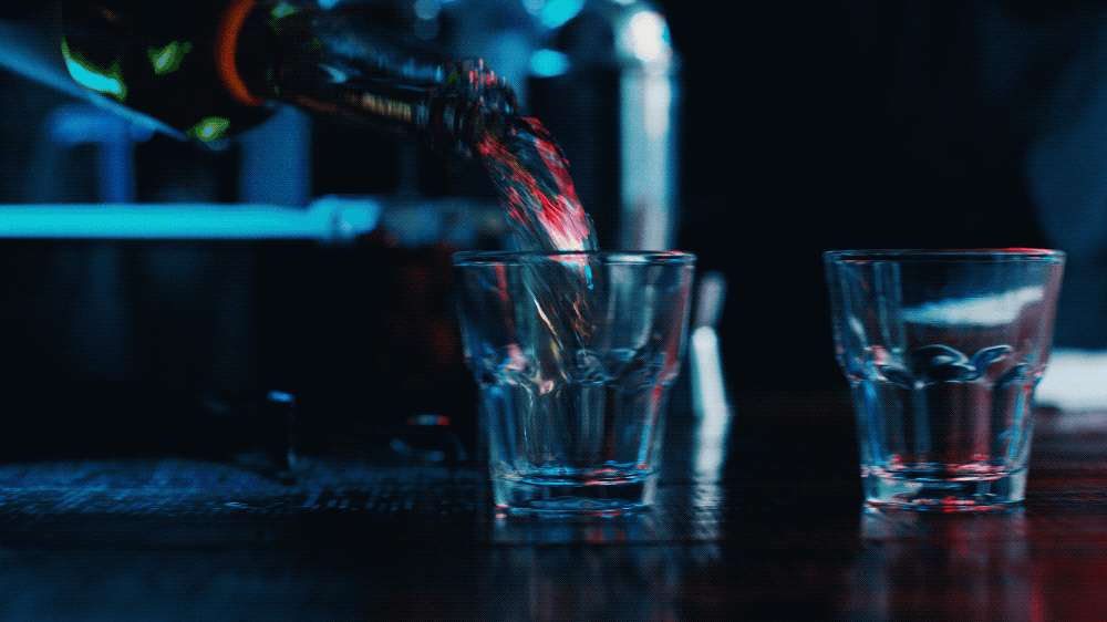 A person pouring a red beverage from a bottle into a glass on a dark table, with another empty glass beside it.