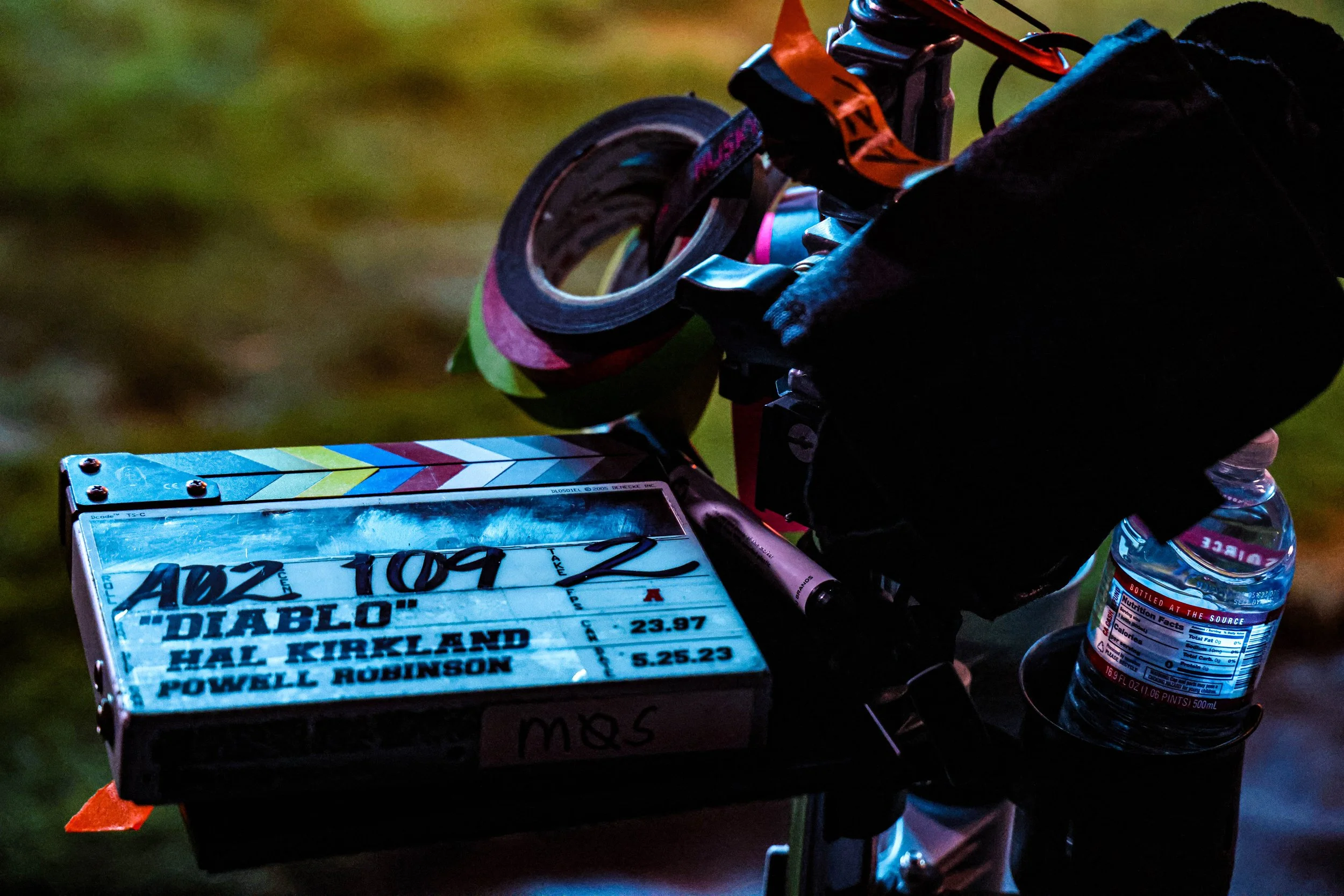 Close-up of a bicycle dashboard with a movie ticket attached, water bottle, black bag, and colorful duct tape on the handlebar.