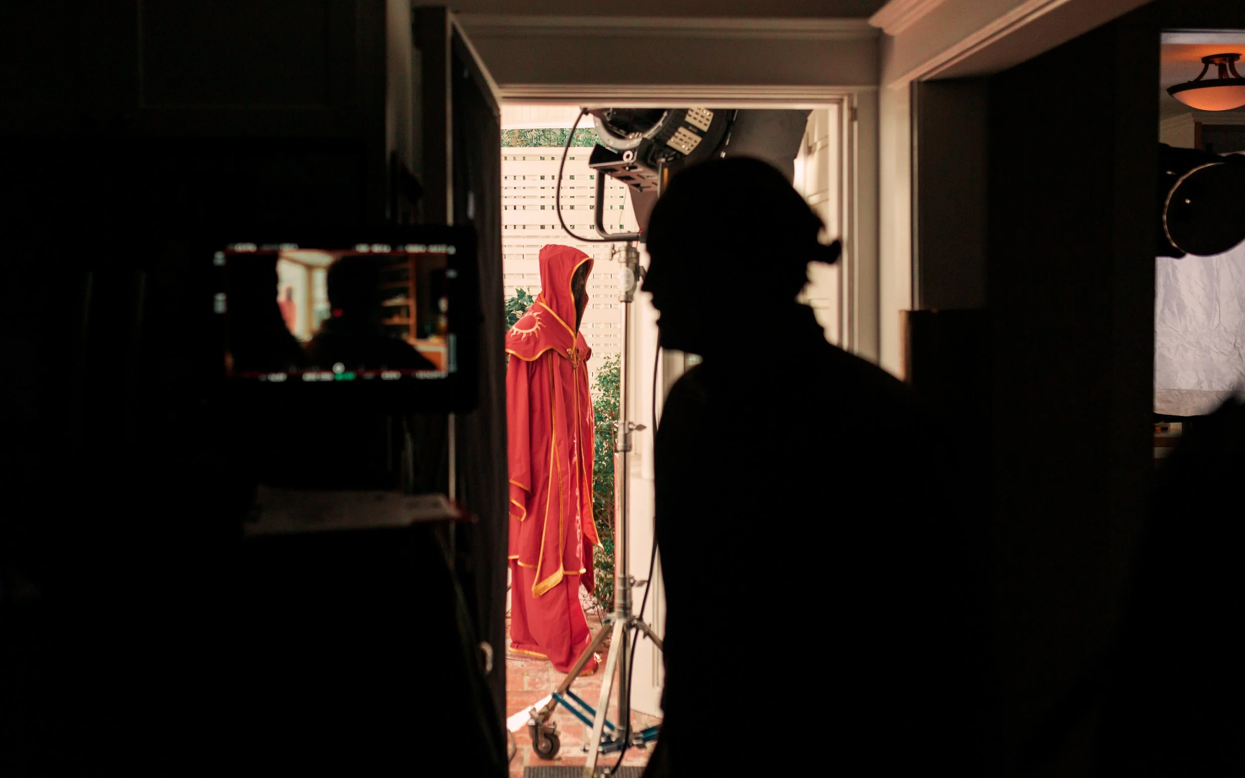 Silhouette of a person in a dimly lit room with a brightly lit doorway leading outside, where a costume or garment with draped fabric and a hood is hanging.