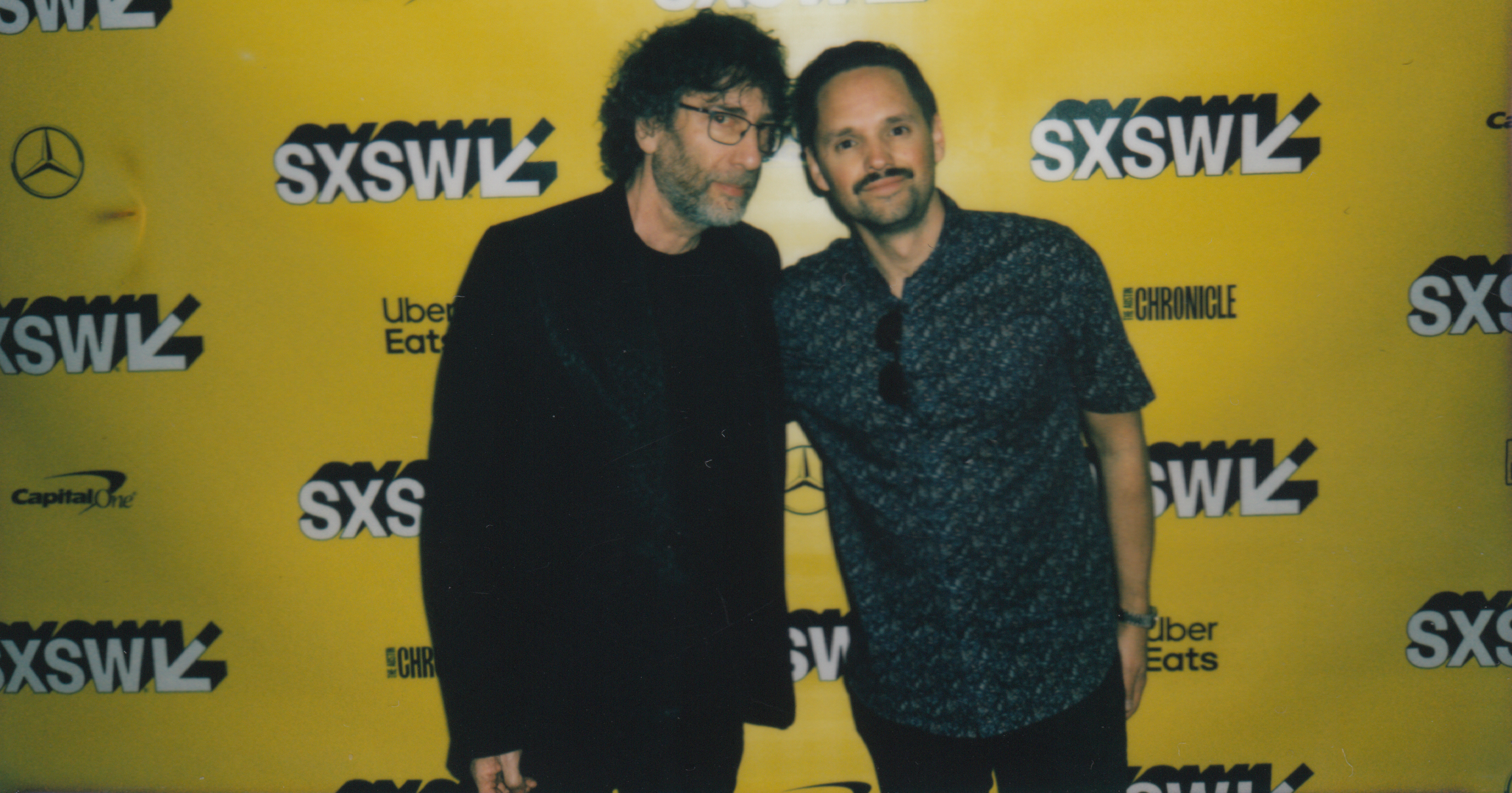 Two men posing together at SXSW event in front of a yellow backdrop with SXSW logos, Uber Eats, Capital One, and The Austin Chronicle branding. Good Omens Amazon Hal Kirkland
