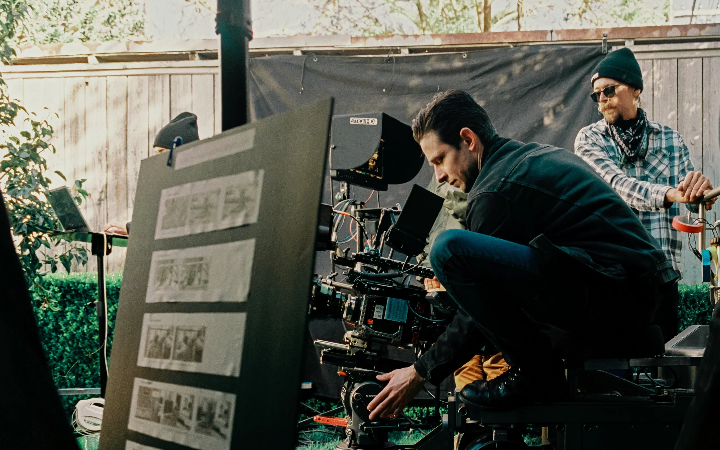 Film crew operating camera equipment outdoors, with one person crouching and adjusting the camera, and another standing with sunglasses and a beanie, in front of a wooden fence.