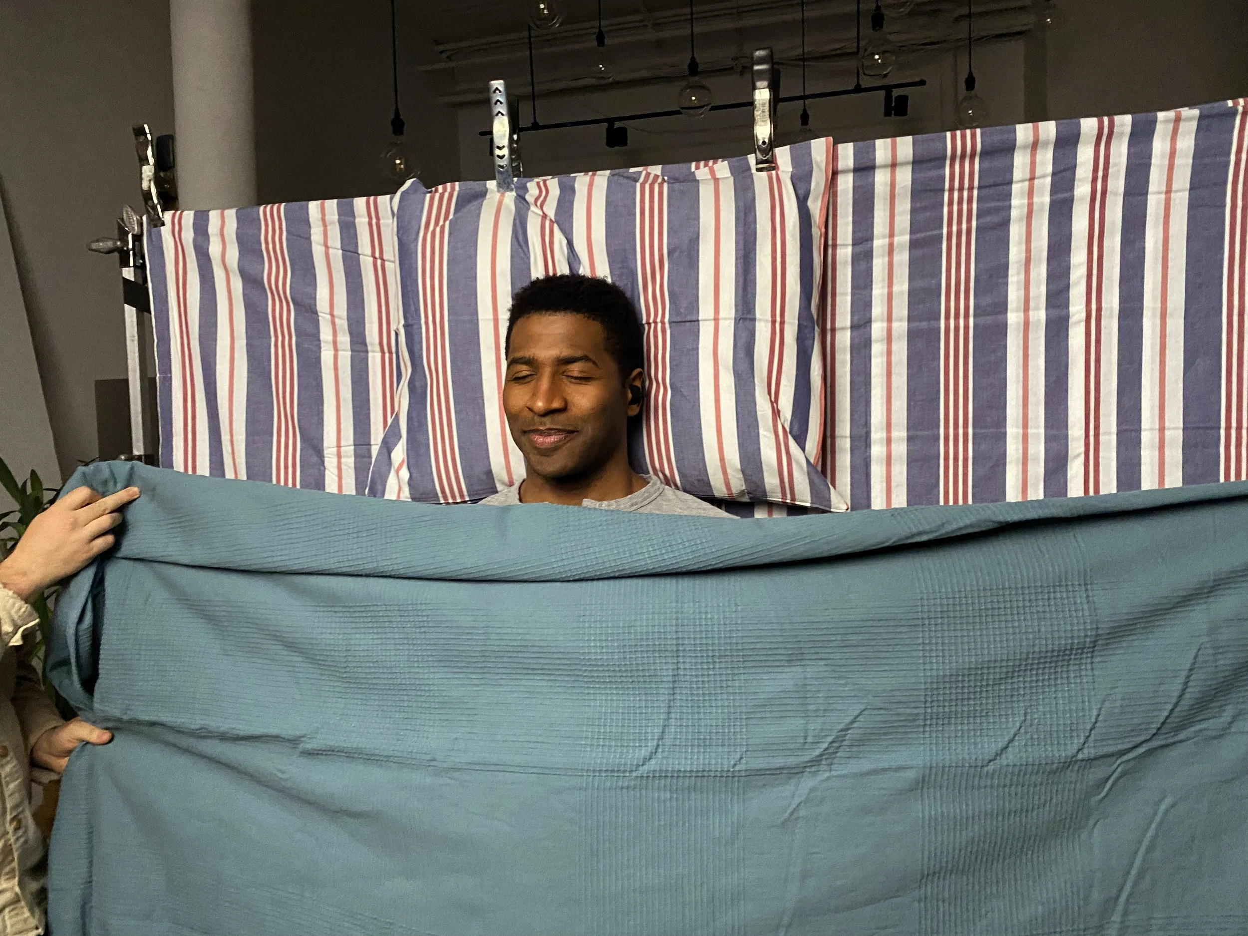 A man with closed eyes, standing behind a blue blanket held by someone off-camera, in a room with a striped backdrop and hanging lights.