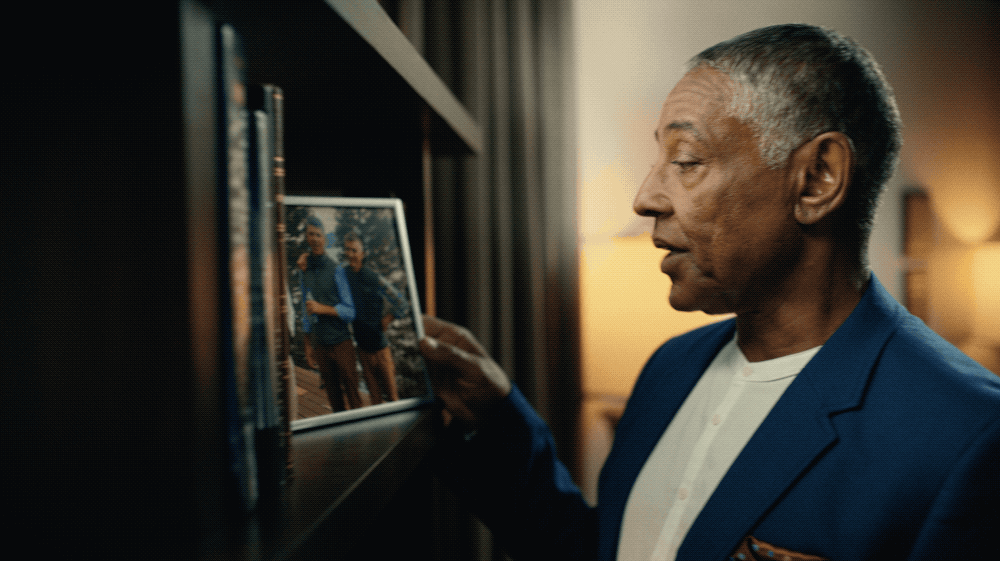 An older man in a blue suit looks at a framed photo on a bookshelf.