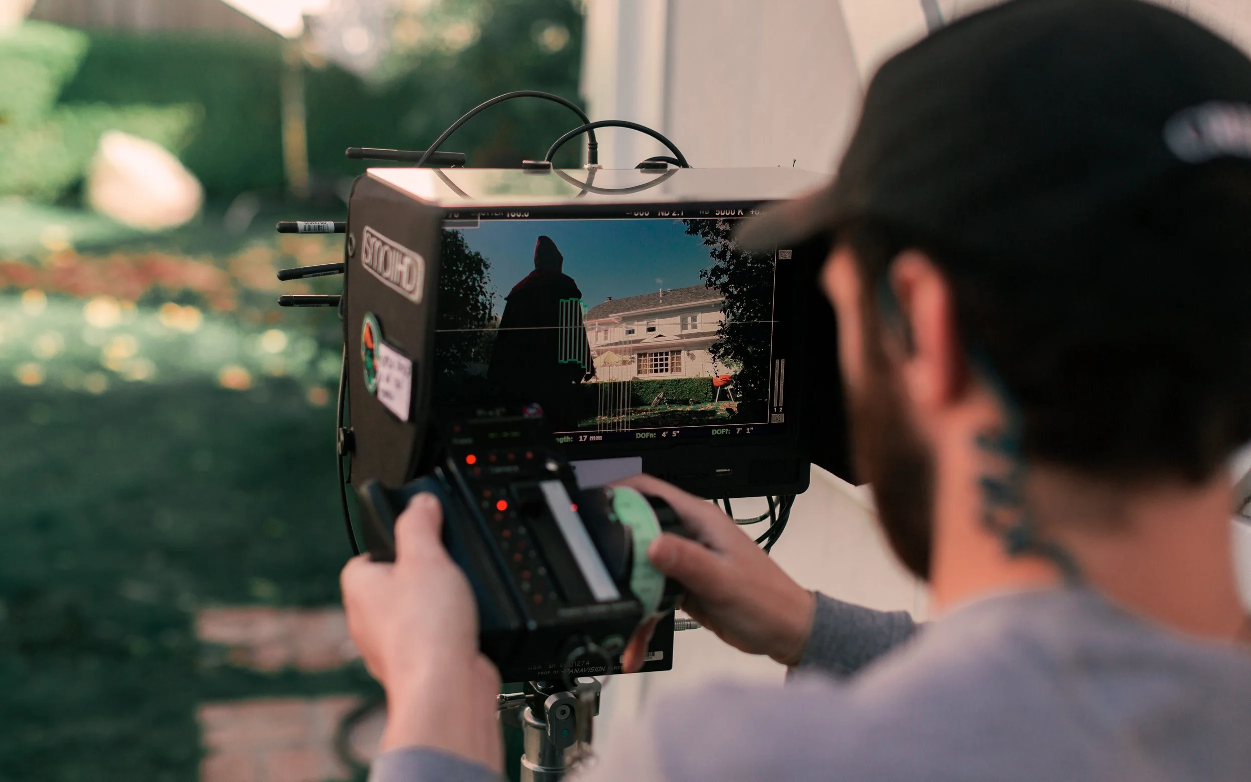 A filmmaker operating a camera on a tripod records a scene showing a person in a hoodie standing outside, in front of a house with greenery.