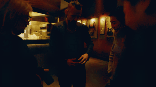 Group of people talking in a dimly lit room, possibly a bar or restaurant, with warm lighting and framed pictures on the wall.