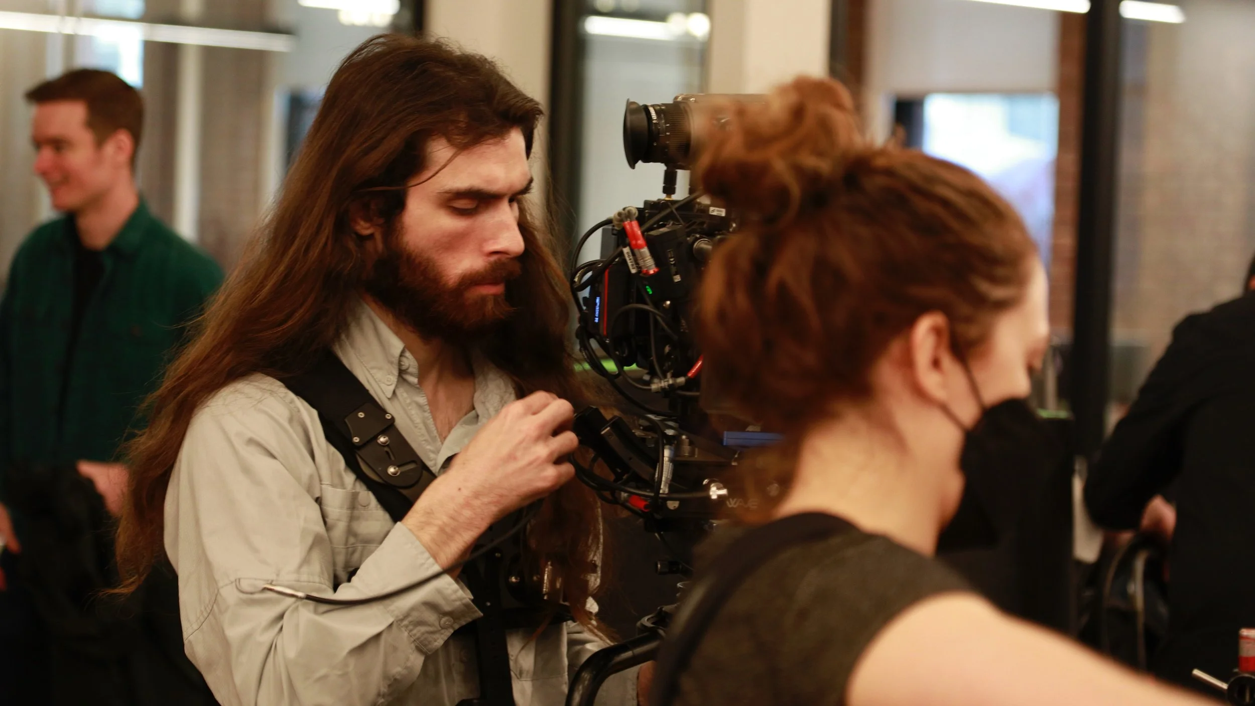 A man with long hair and a beard operating a professional film camera, with a woman wearing a face mask standing nearby. In the background, another man is smiling and looking away.