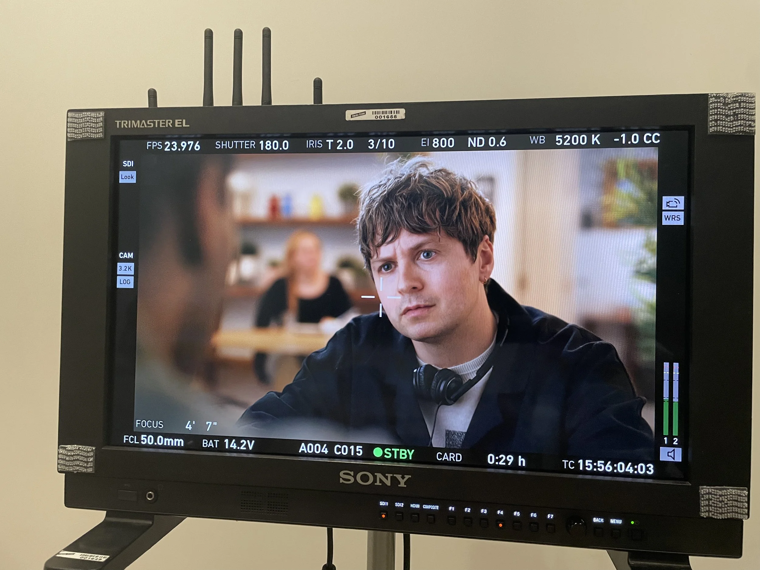 A person with brown hair and blue eyes, wearing headphones, is sitting in front of a camera on a production monitor, with a blurred background featuring a woman sitting at a table.