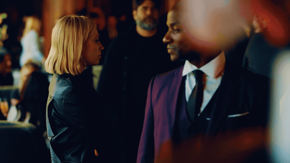 Two people, a woman and a man, are having a serious conversation at a formal event. The woman has blonde hair and wears a black jacket. The man has dark skin, is smiling, and wears a suit with a tie.