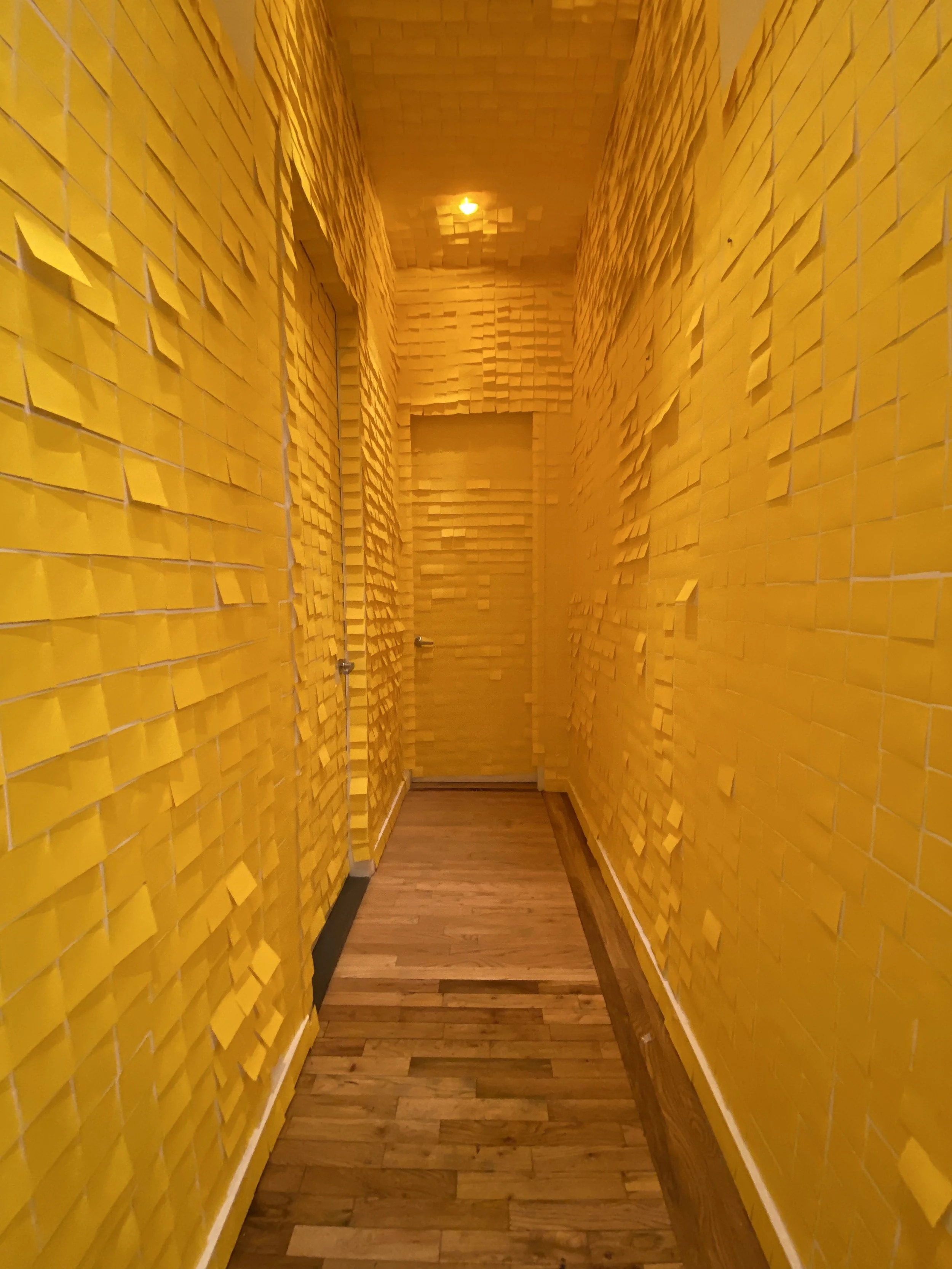 A hallway with walls and ceiling covered in numerous yellow sticky notes, with a wooden floor and a door at the end.