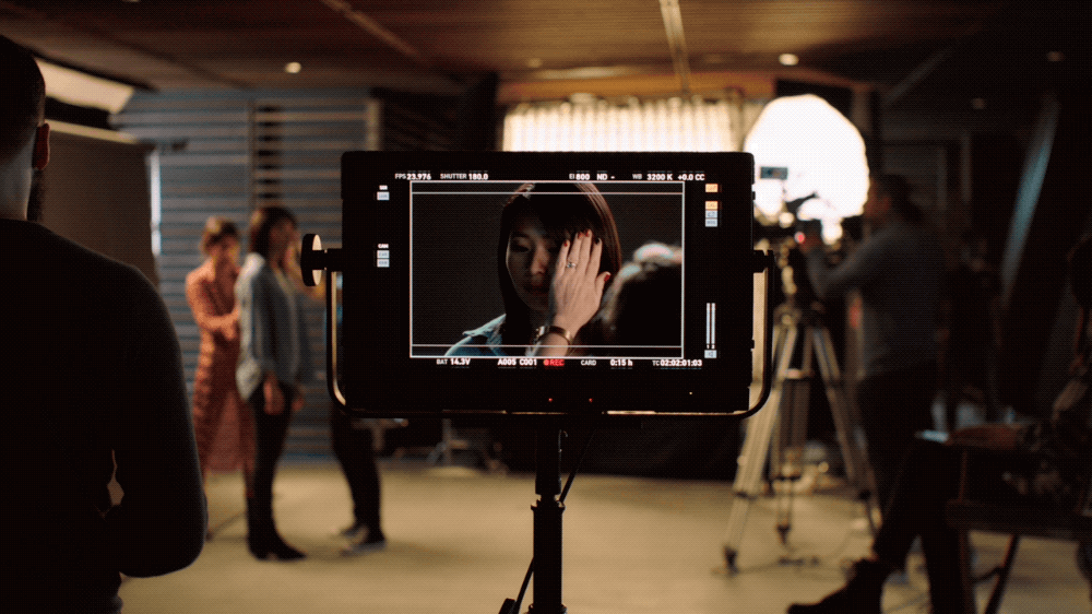 A woman is being filmed on a professional video camera, with a scene of three women standing and talking in the background.