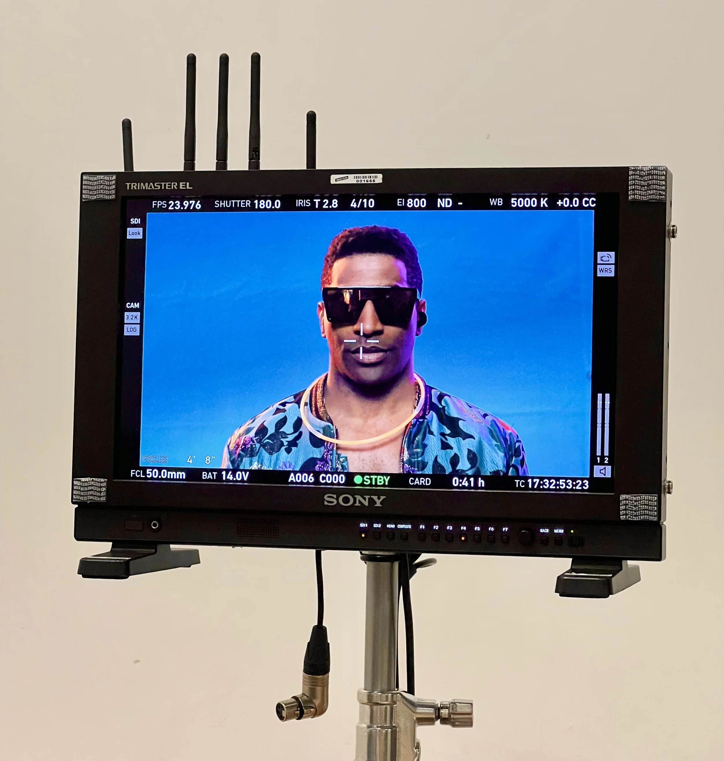 A professional video monitor displaying a portrait of a man with dark skin, wearing sunglasses, a colorful patterned shirt, and a gold chain, set against a blue background.
