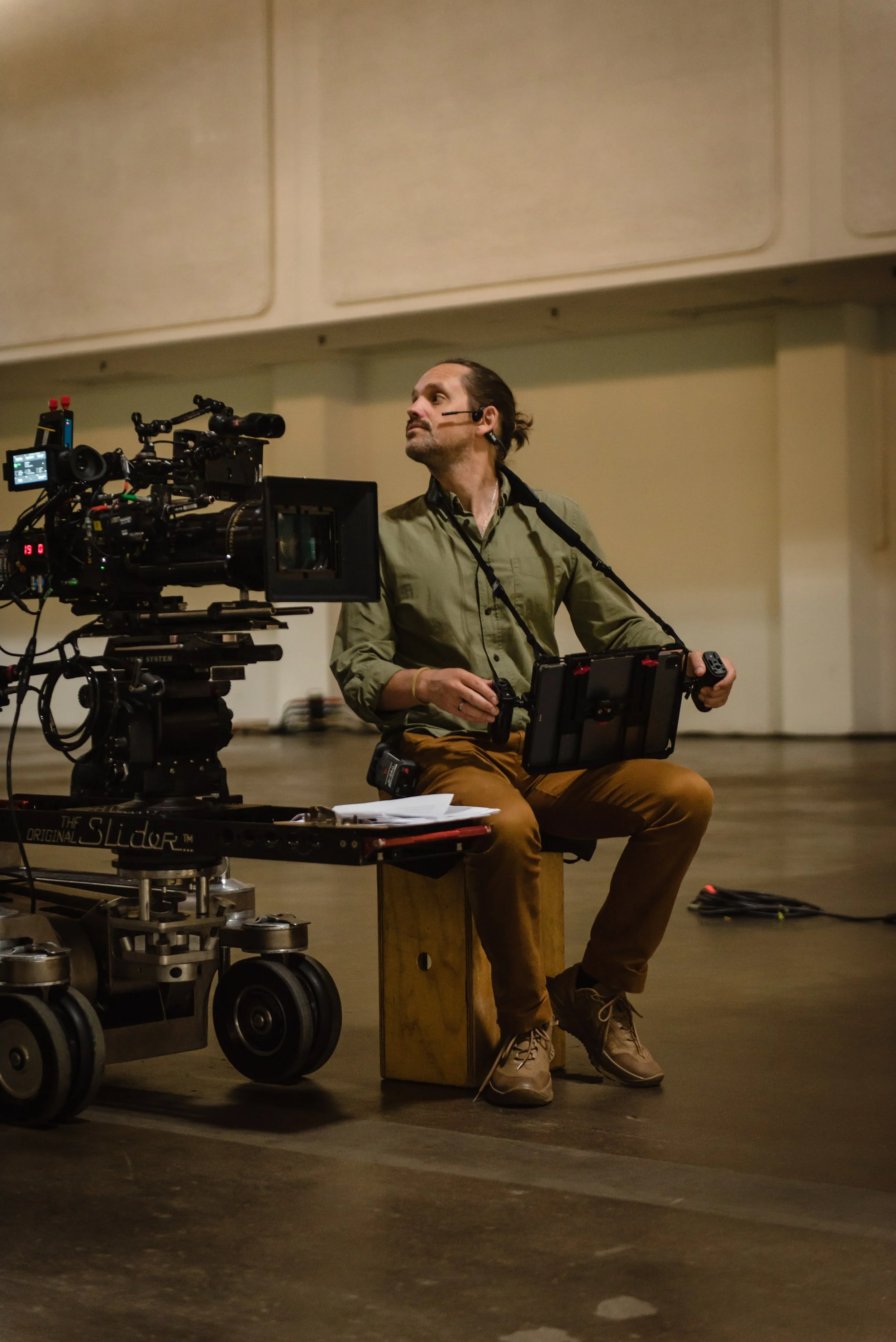 A man operating a professional film camera on a dolly, sitting on a small wooden stool inside a large indoor space.