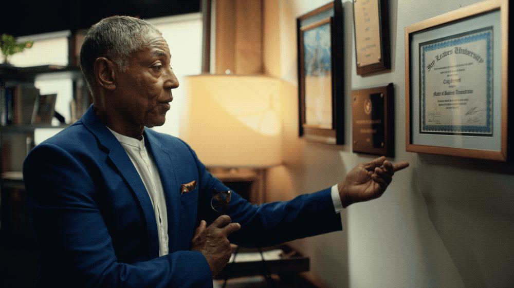 An older man in a blue suit is pointing at a framed certificate on a wall in an office or library. The room has warm lighting and shelves with books.