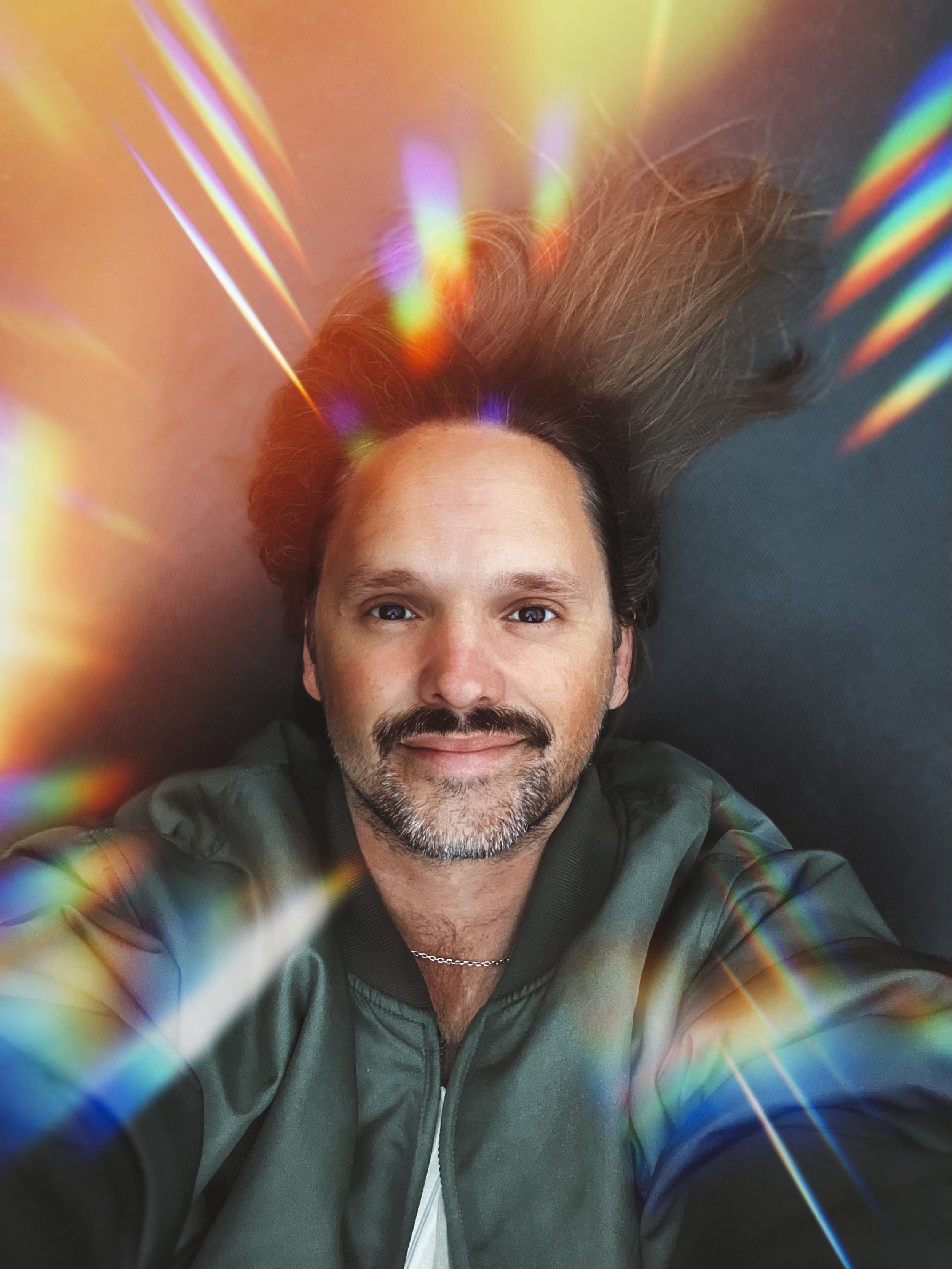 A man with brown hair and a beard lying on a black surface, smiling, with colorful light reflections and rainbow streaks around him.