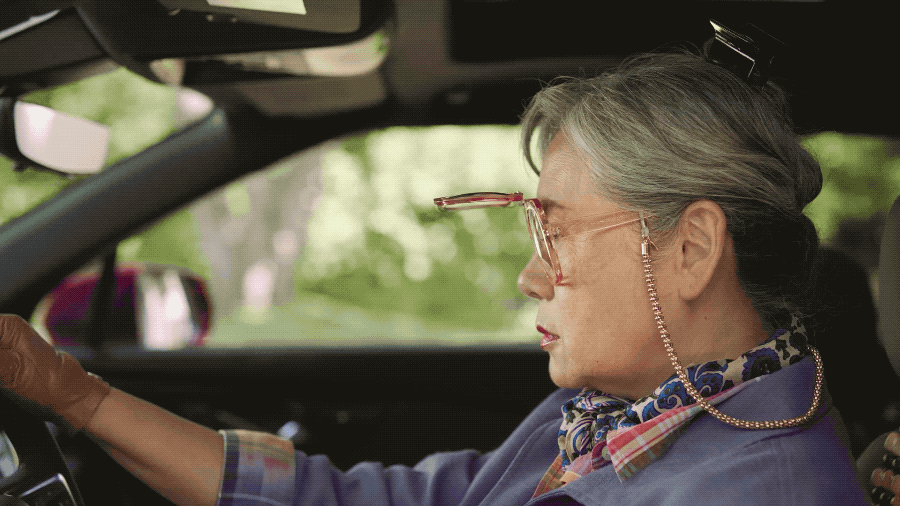 An elderly woman with gray hair styled in a bun, wearing glasses with a chain, a blue plaid scarf, and a blue jacket, driving a car.